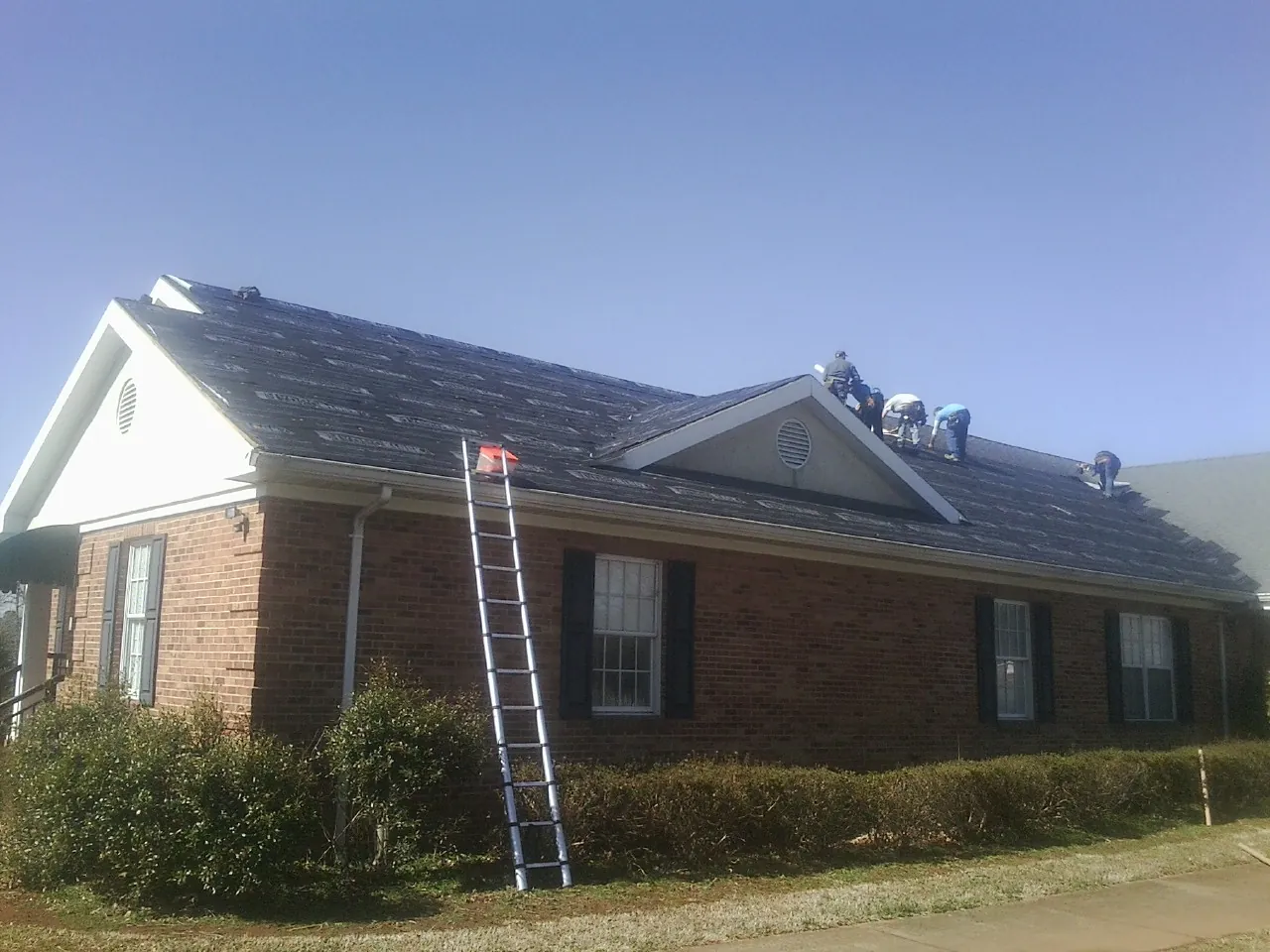 Crew tearing shingles off a brick ranch roof with a ladder set — Metal Roof Repair work by our crew in Bevington