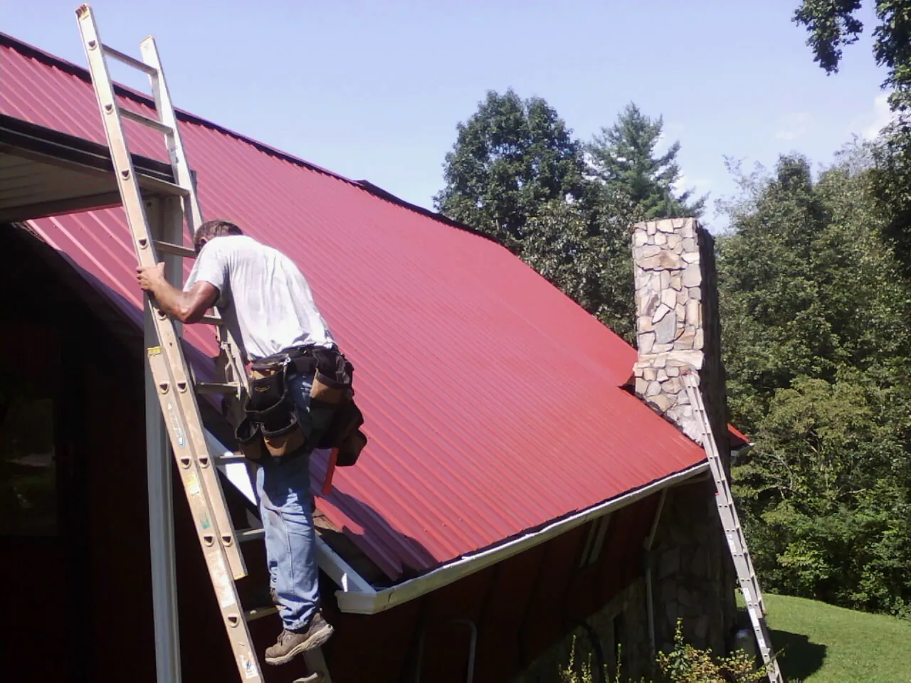Roofer on a ladder beside a red metal roof and stone chimney — Soffit & Fascia Repair by Roofing Brothers in Bevington